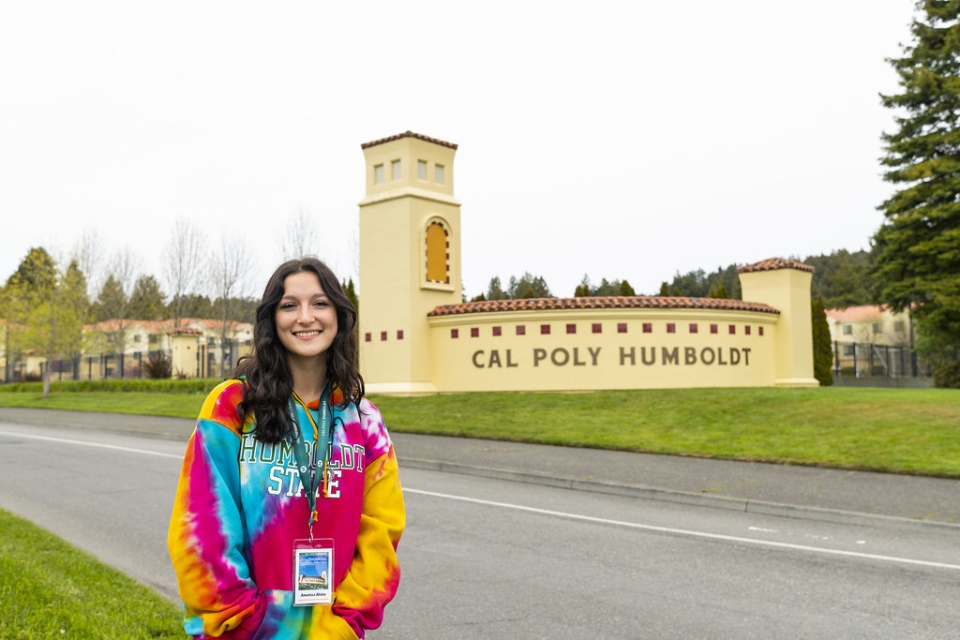 A photo of someone standing and smiling in front of the Cal Poly Humboldt university sign